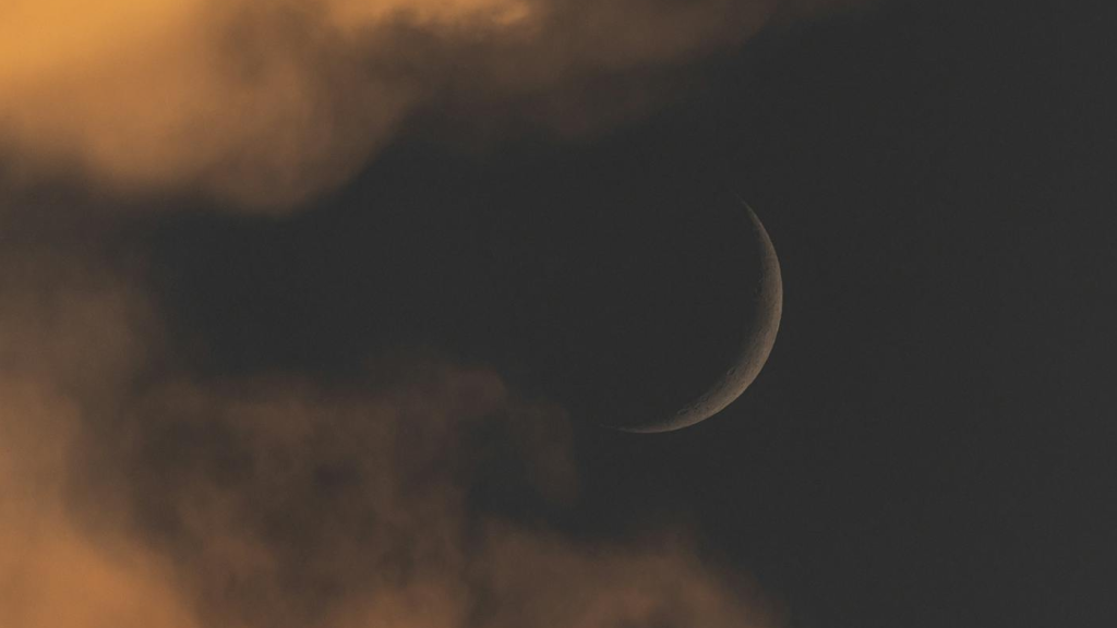 Тънък лунен сърп (Новолуние) над тъмно небе с облаци в оранжев и кафяв тон